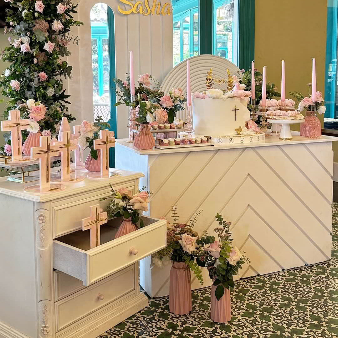 A decorated event altar setup with a white chevron counter, pink floral arrangements, candles, wooden crosses, and a white cake, set in front of a green-framed glass door with a floral arch reading 'Sasha'.