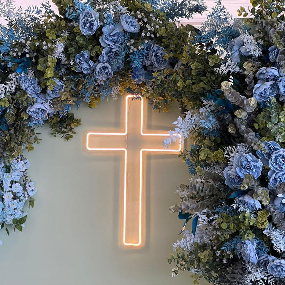 Neon cross light surrounded by decorative floral arrangement on a plain background