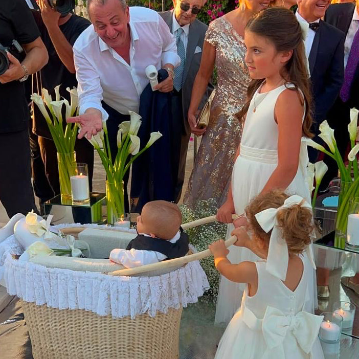 A baby in a white outfit sits in a woven bassinet with a white ruffled liner, surrounded by guests at an outdoor event. A young girl in a white dress with a large bow stands nearby, and flowers and greenery decorate the background.