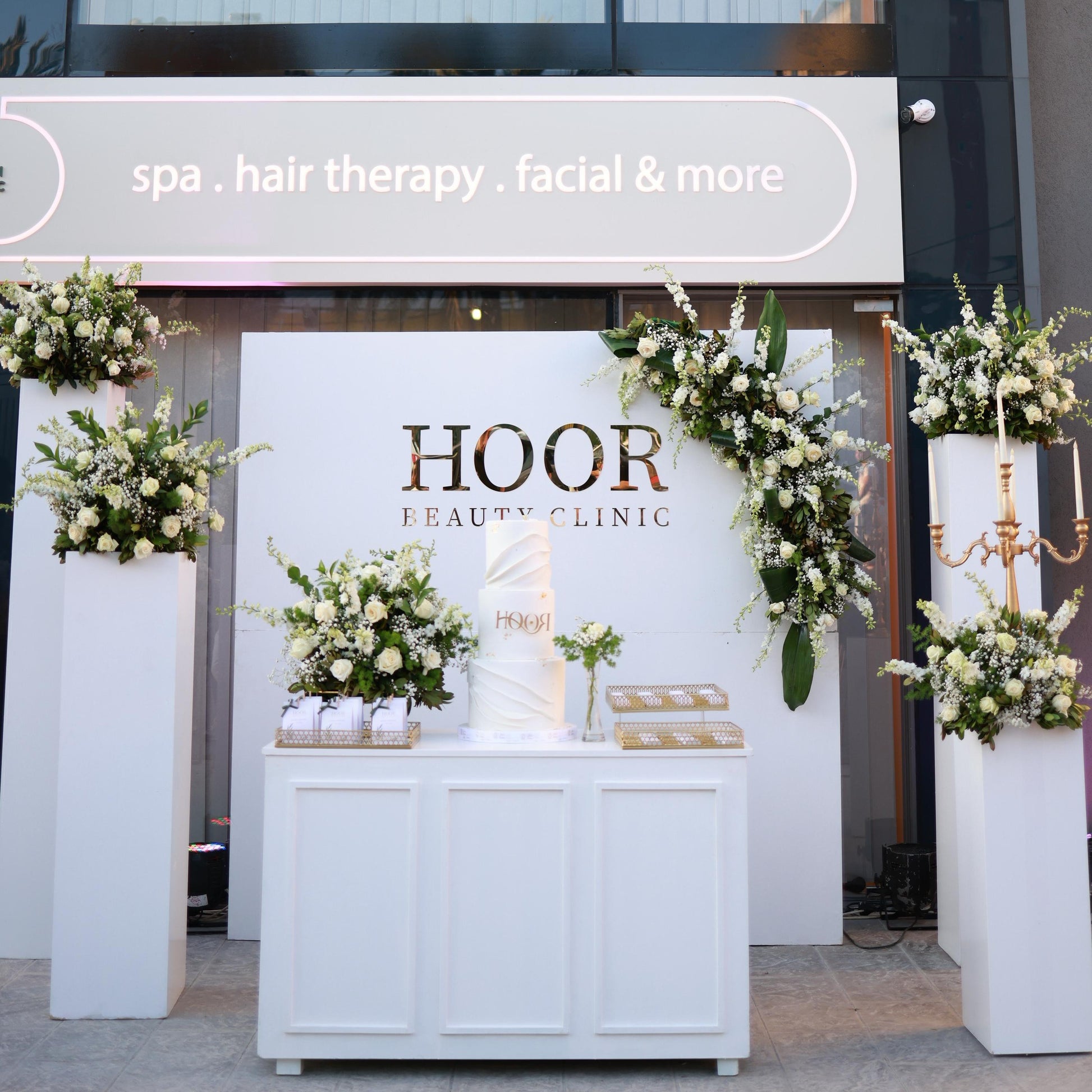 A white reception counter and backdrop for Hoor Beauty Clinic, decorated with white floral arrangements and greenery, set in front of a glass storefront with a sign reading 'spa . hair therapy .