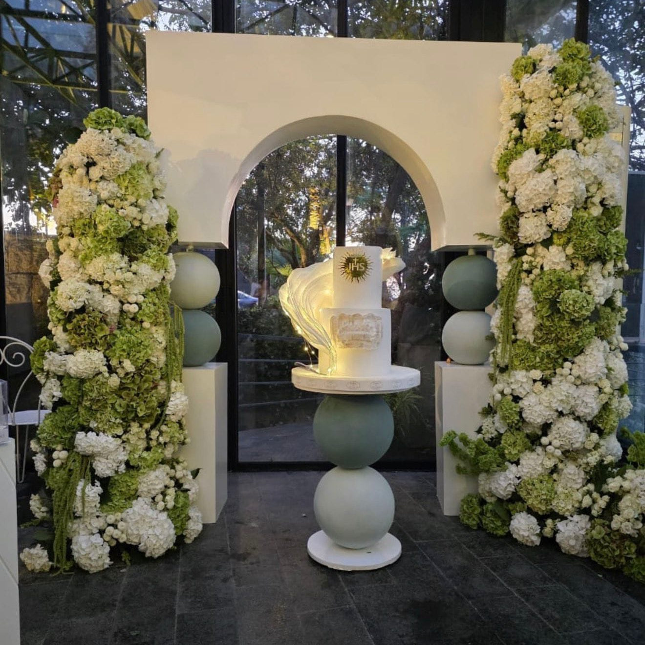 A decorative wedding-style floral installation with two tall columns of white and green flowers flanking an arched white backdrop, with a tiered white cake displayed on a round pedestal in the center.