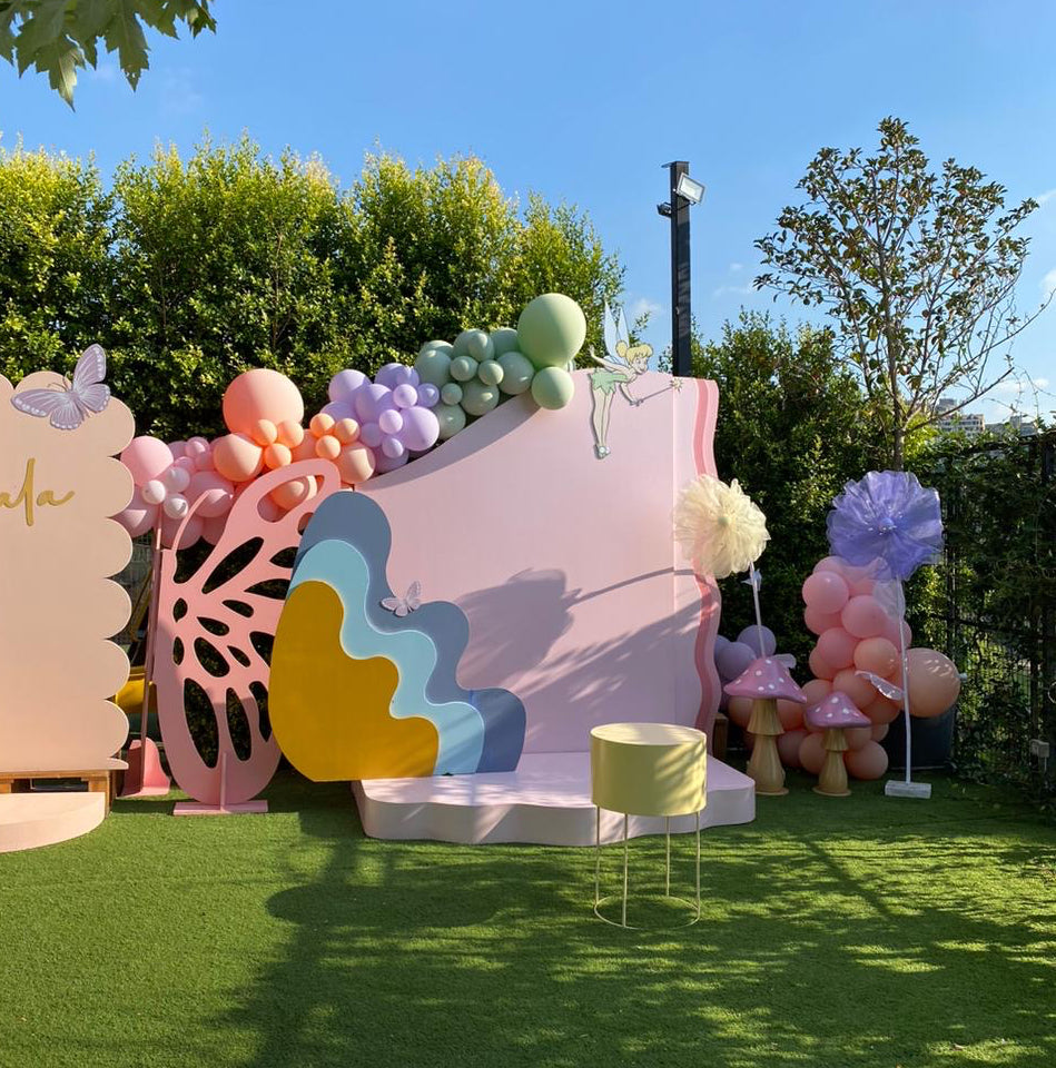 Outdoor party backdrop setup on green grass with a pastel pink scalloped board, colorful balloon garlands, decorative paper cutouts, a small yellow stool, and trees in the background under a blue sky.