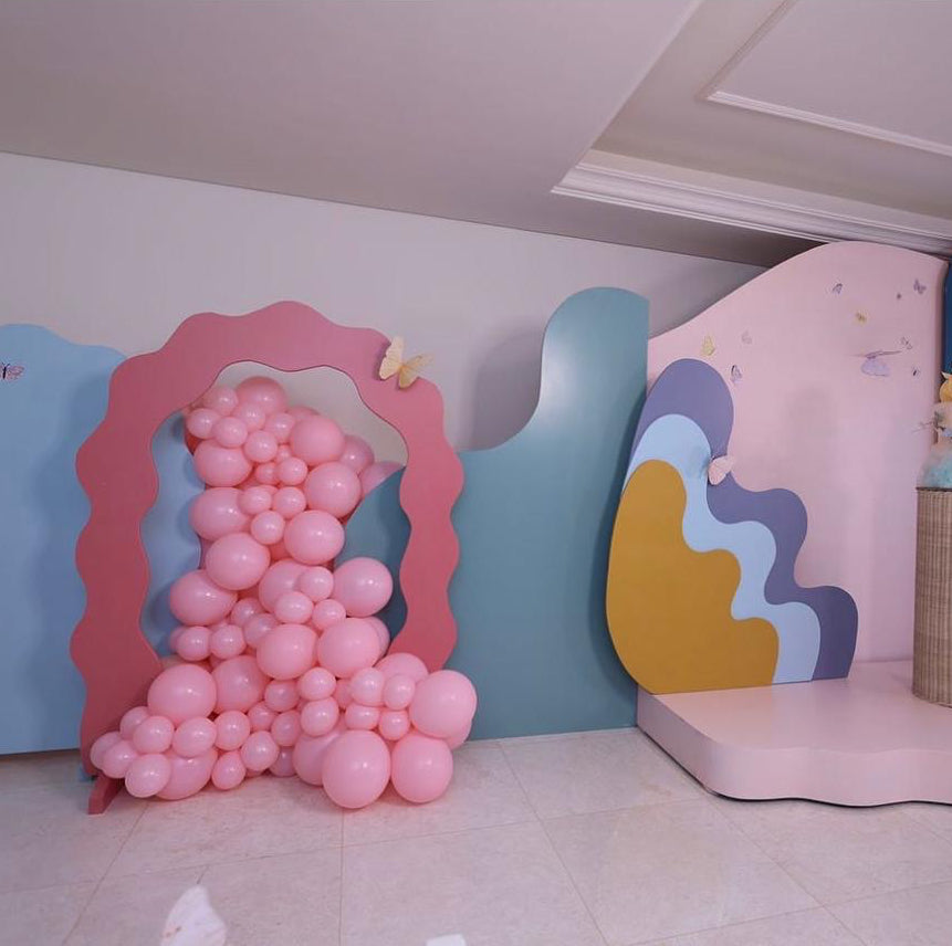 A pastel party backdrop setup in a room, featuring wavy cutout panels in pink, blue, yellow, and lavender, with a cluster of pink balloons arranged inside one frame-shaped panel and a decorative pillar on a pink platform.