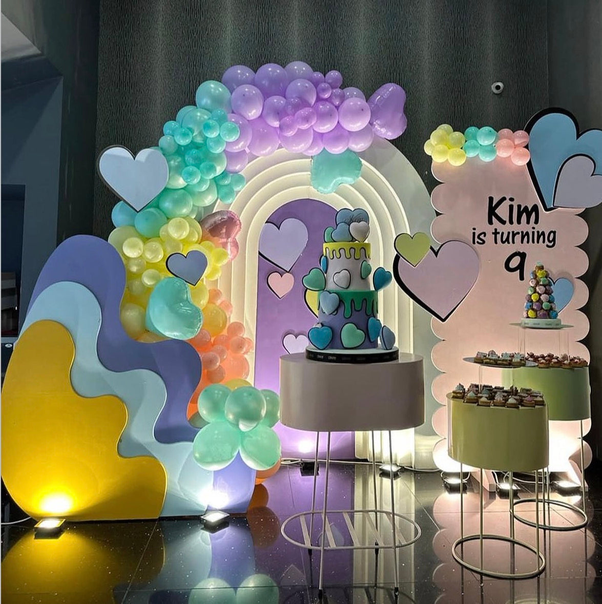 A colorful birthday party backdrop setup with a rainbow arch made of illuminated balloons, pastel abstract wall panels, heart decorations, and a dessert table with cupcakes. A large sign reads 'Kim is turning 9'.