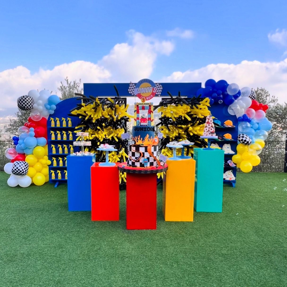 Colorful birthday setup with balloons, cake, and decorations against a blue sky.