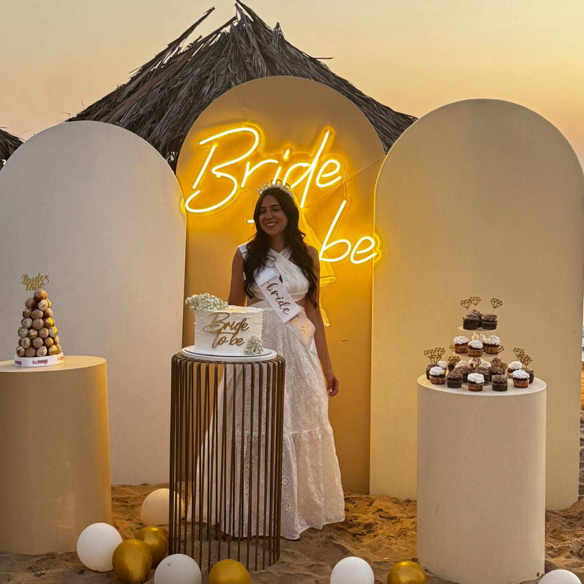 Woman in a white dress standing in front of a 'Bride to be' neon sign with dessert display.