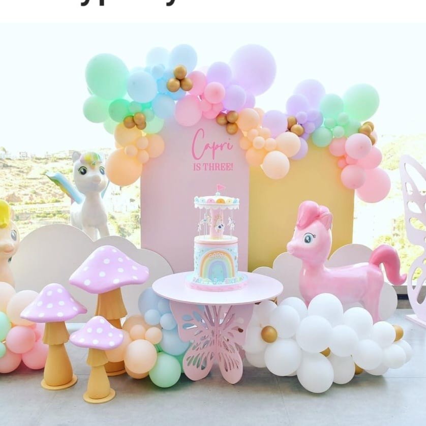 Pastel-coloured balloon arch with a unicorn, cake, and pink pony toy on a table for a girl birthday