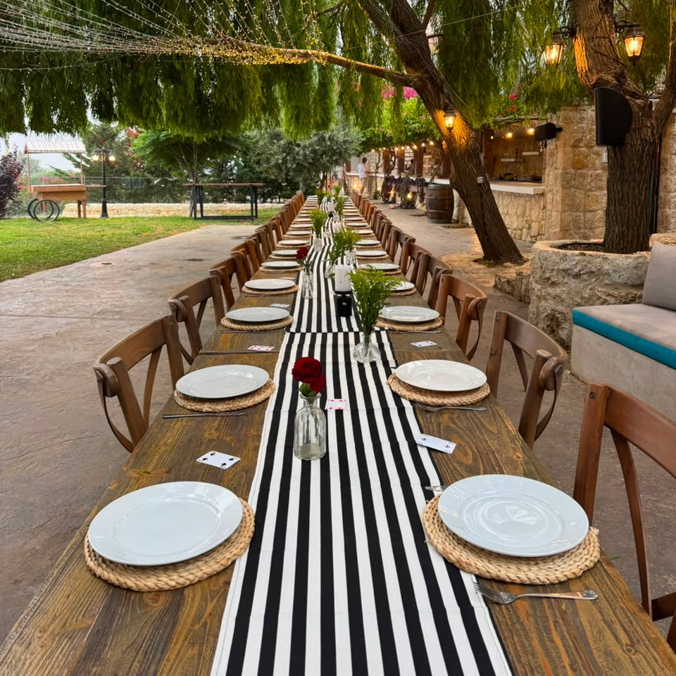 An outdoor dining setup with a long wooden table set with white plates, woven placemats, a black-and-white striped runner, and small floral centerpieces under weeping trees with string lights.