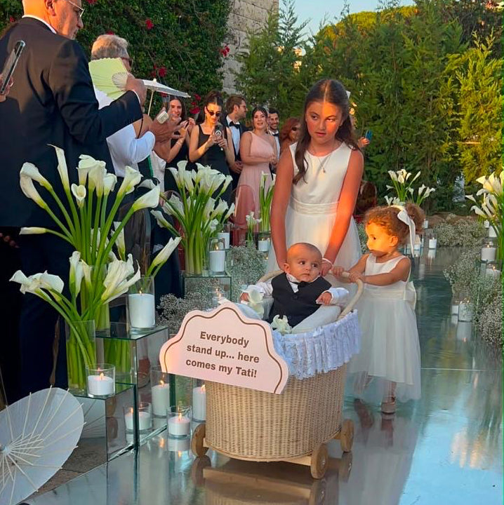 A woman in a white sleeveless dress pushes a wicker basket stroller at an outdoor wedding ceremony. A young girl in a white dress stands beside the stroller, and guests are seated in the background.