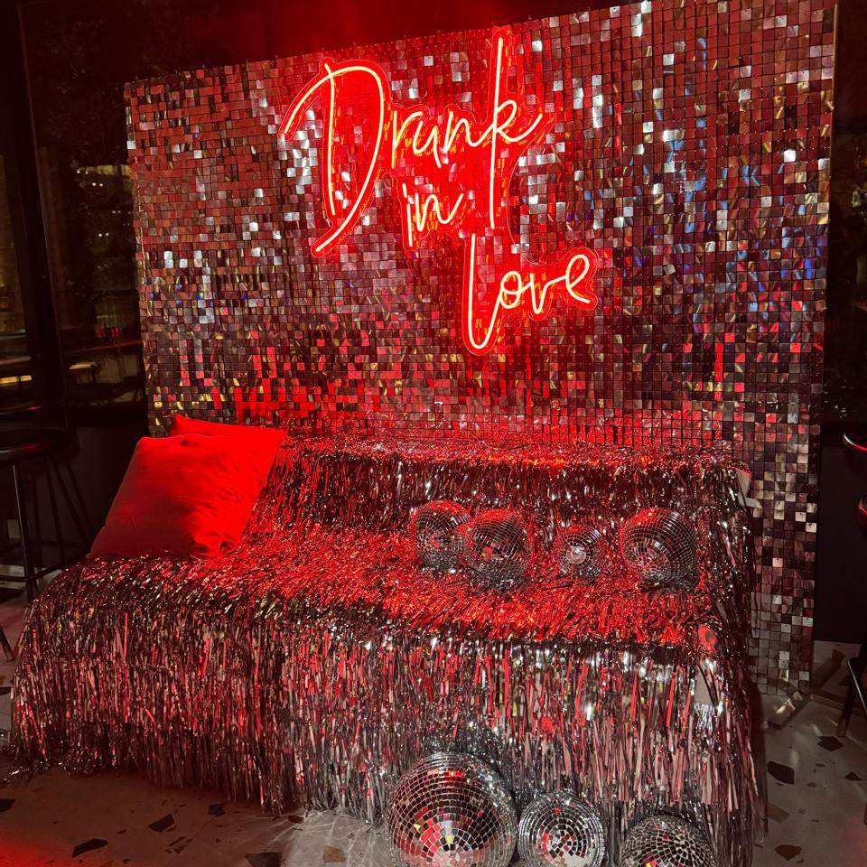 Decorative setup with a glittery backdrop, disco balls, and a neon sign reading 'Drunk in Love' for a bachelorette party.