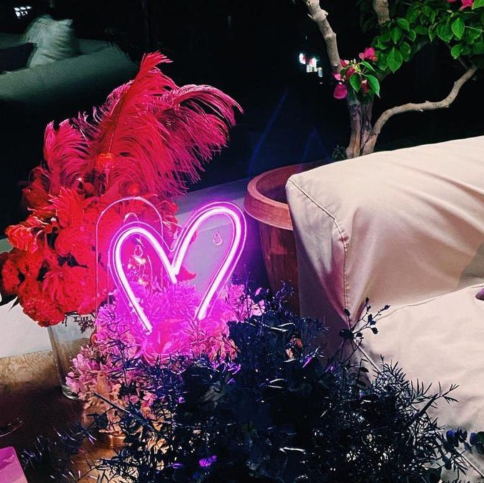 Neon heart light with pink feathers and flowers on a table at night.