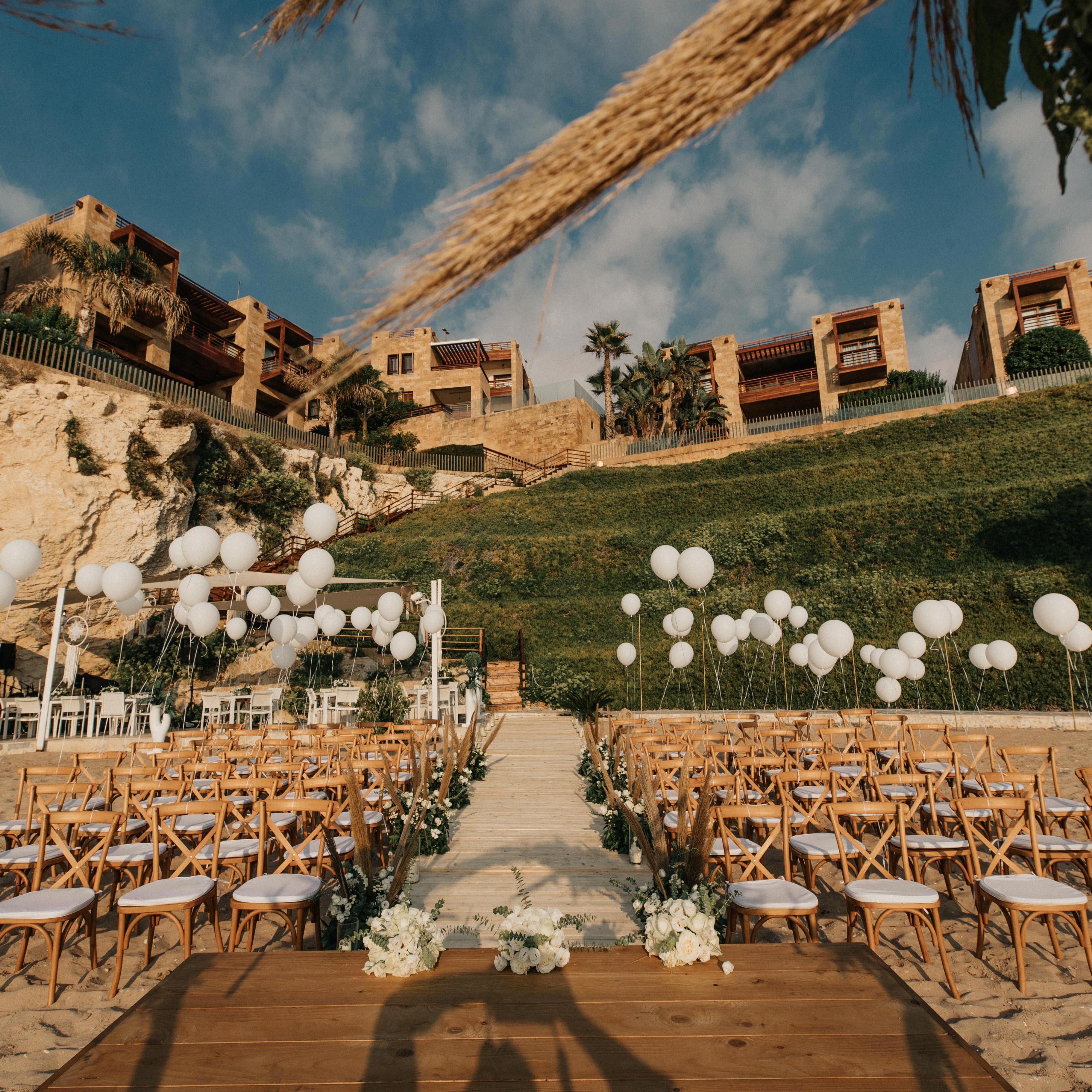 Outdoor wedding setup with chairs, flowers, and balloons on a beach with buildings in the background.