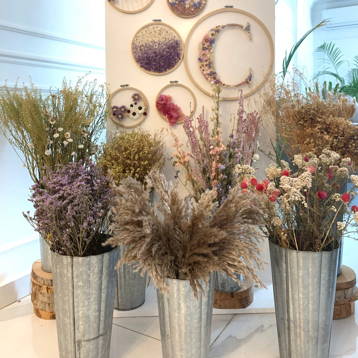 A styled display of dried flowers and botanicals arranged in galvanized metal vases in front of a white wall sign that reads 'Capri's Flower Shop.' The display includes purple lavender, beige pampas grass, cream flowers, and small pink and red blooms, with circular hoop art and pressed floral pieces mounted on the wall.