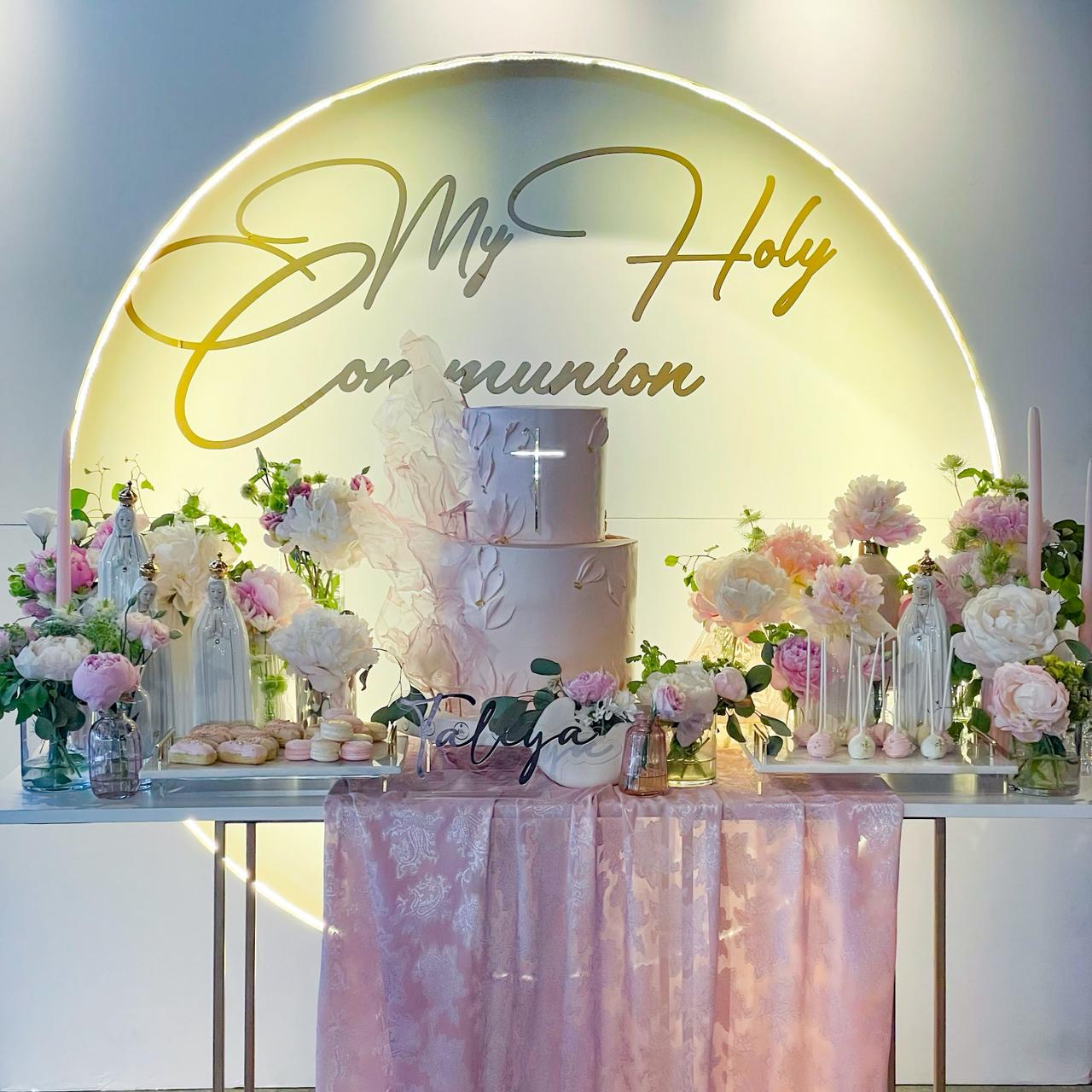 Decorative table setup for a religious celebration with flowers and a cake, featuring 'My Holy Communion' text.