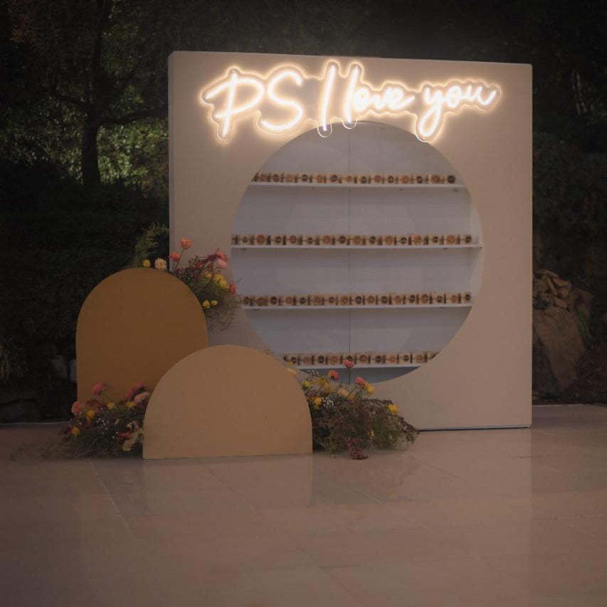 Neon sign with 'PS I love you' on a white board with floral decorations.
