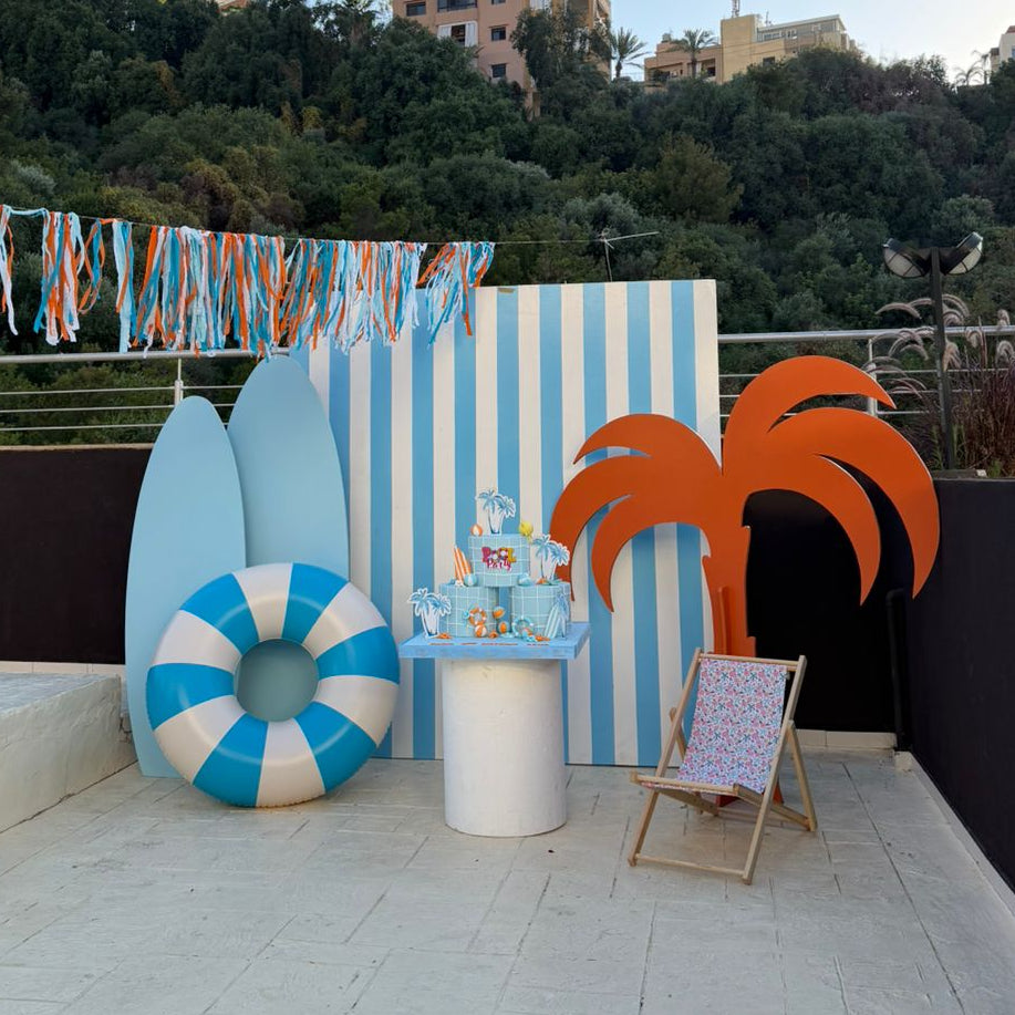 A beach-themed party setup on an outdoor terrace, featuring a blue and white striped backdrop, a blue and white striped inflatable ring, light blue surfboard cutouts, orange palm tree cutouts, and a small decorated table in the center.