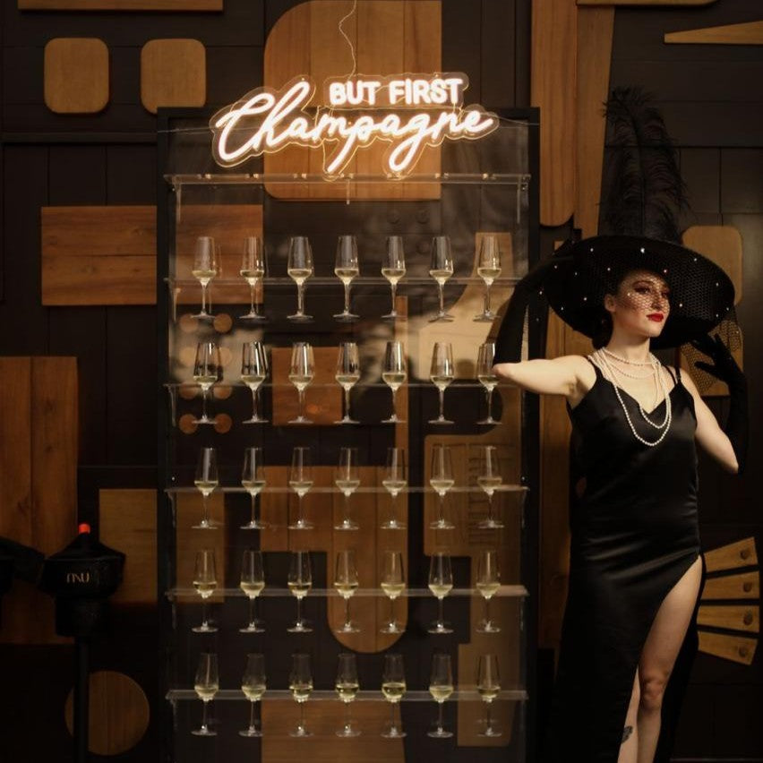 Woman in a black dress and hat standing next to a wall of champagne glasses with 'But First, Champagne' sign.