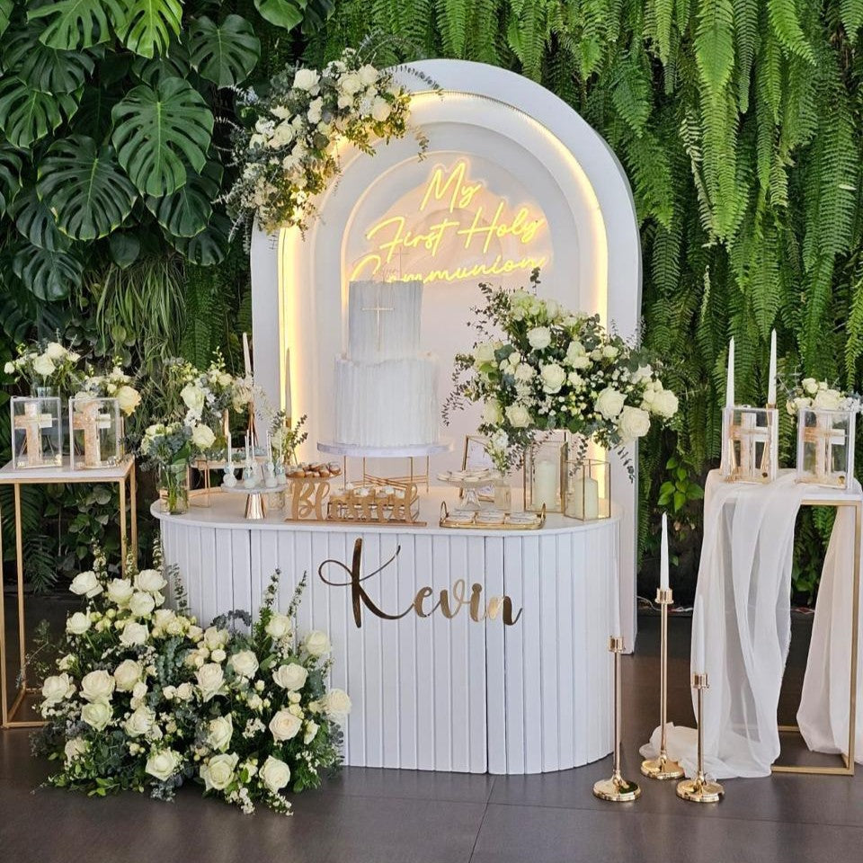 Decorative setup with floral arrangements, a neon sign, and a nameplate against a green leafy background.