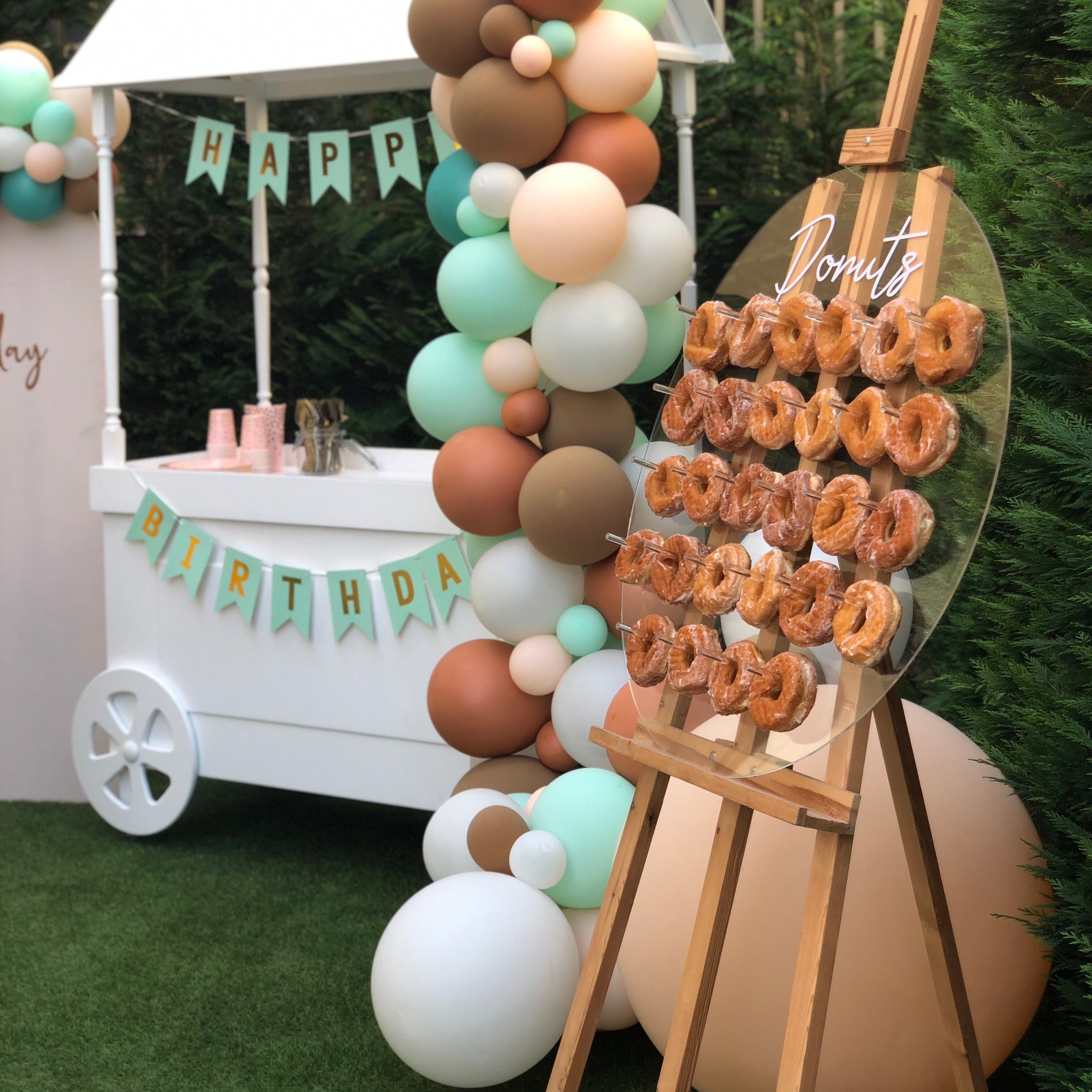 Decorative balloon arch with a 'Happy Birthday' banner and donut display stand outdoors.