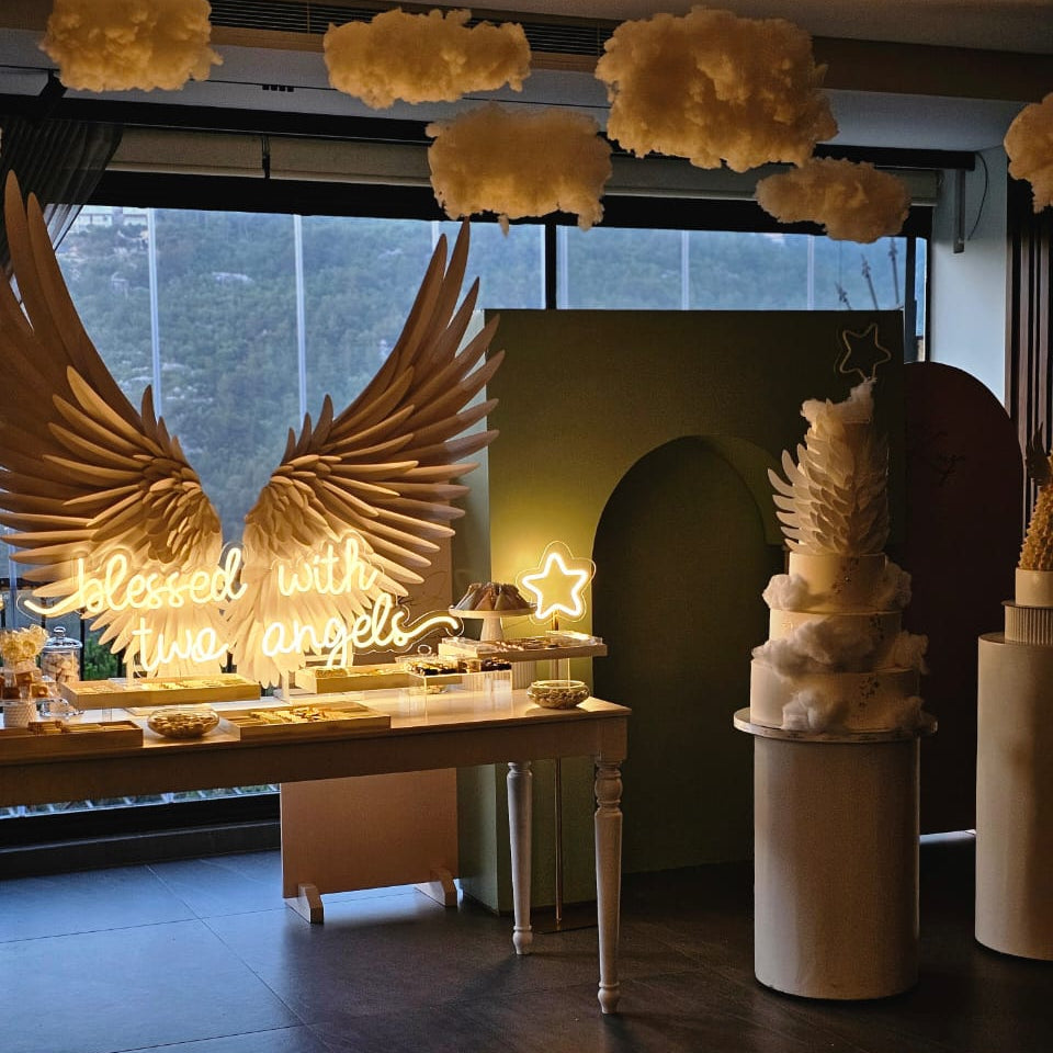 Decorative setup with angel wings, cake, and 'Blessed with little angels' sign in a room.