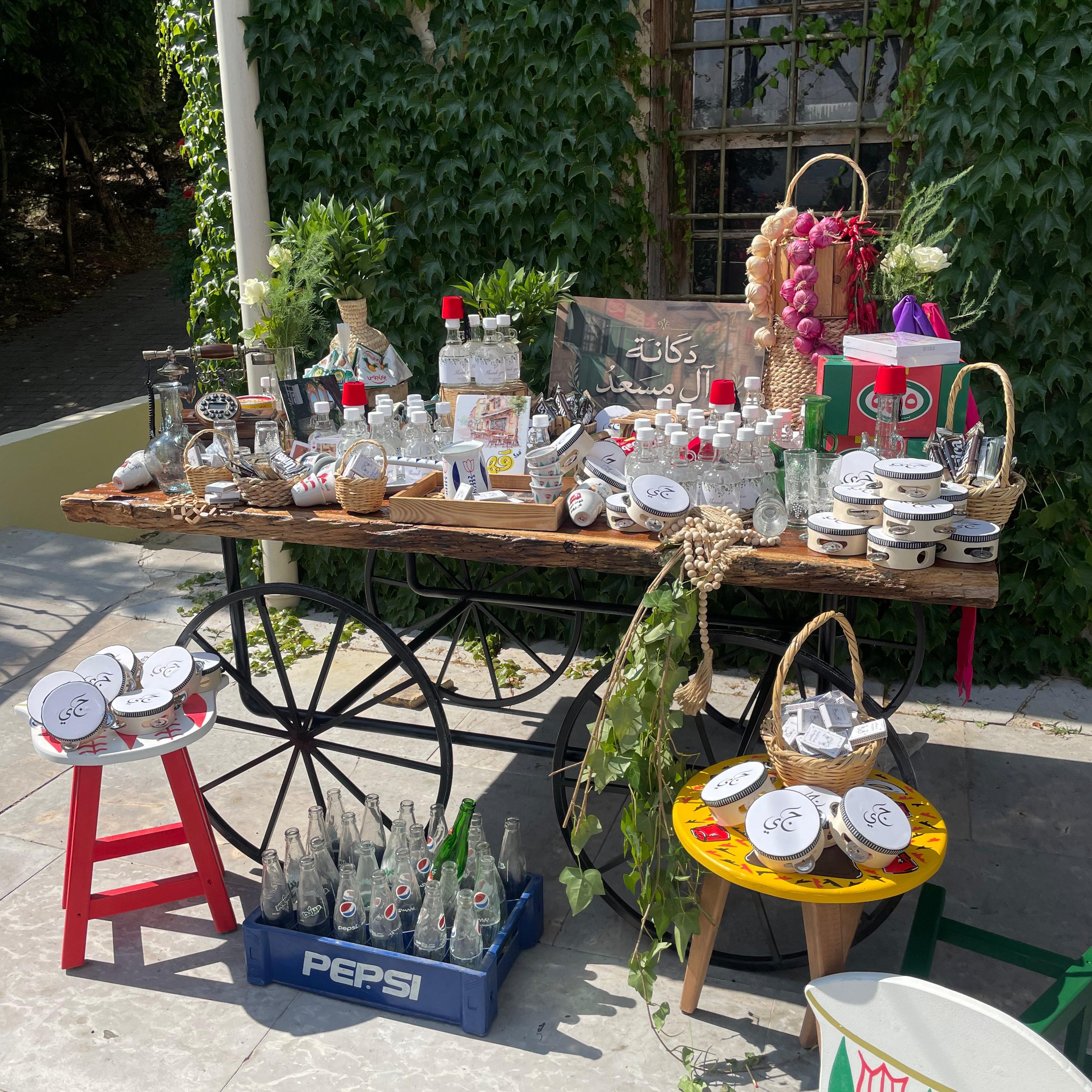 A rustic outdoor display table set up on a patio, covered with numerous small glass bottles, jars, and decorative items. The table is mounted on a vintage black metal wagon wheel, with colorful stools and baskets holding additional products.