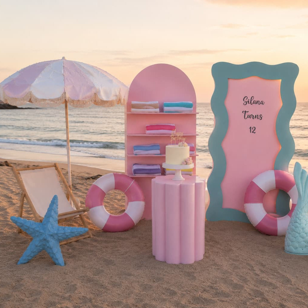 A beach-themed party setup on sand at sunset, featuring a pink and teal scalloped backdrop, a pink shelf with folded towels, a small cake stand, a pink-and-white striped umbrella, lifebuoys, a blue starfish, a teal seashell, and a teal mermaid tail decoration.