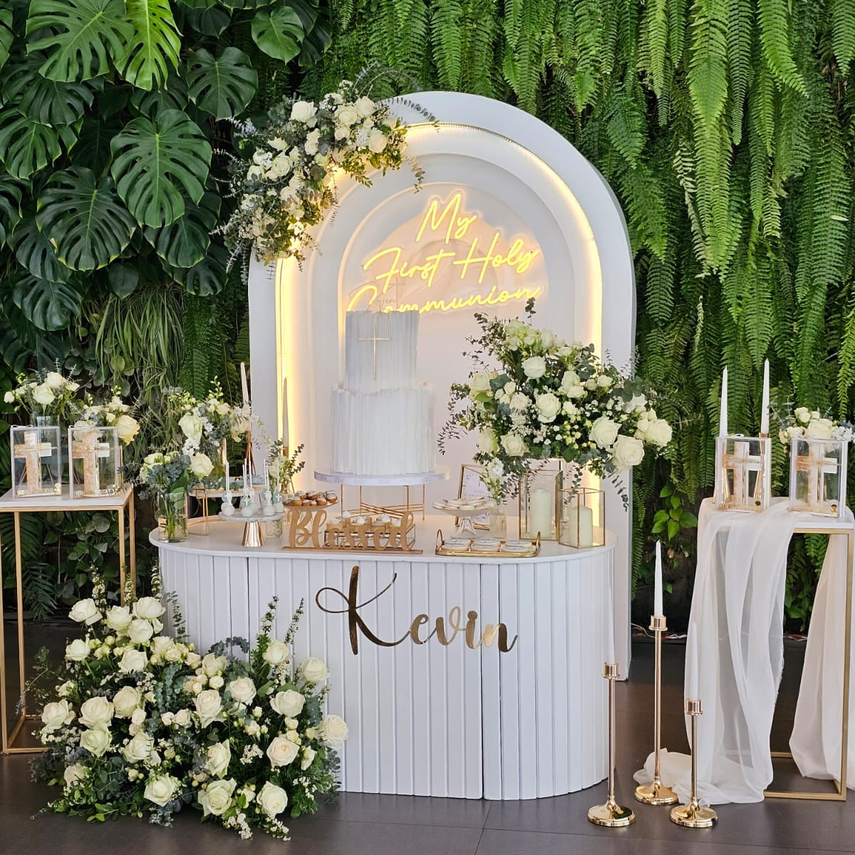 Decorative setup with floral arrangements, a neon sign, and a nameplate against a green leafy background.