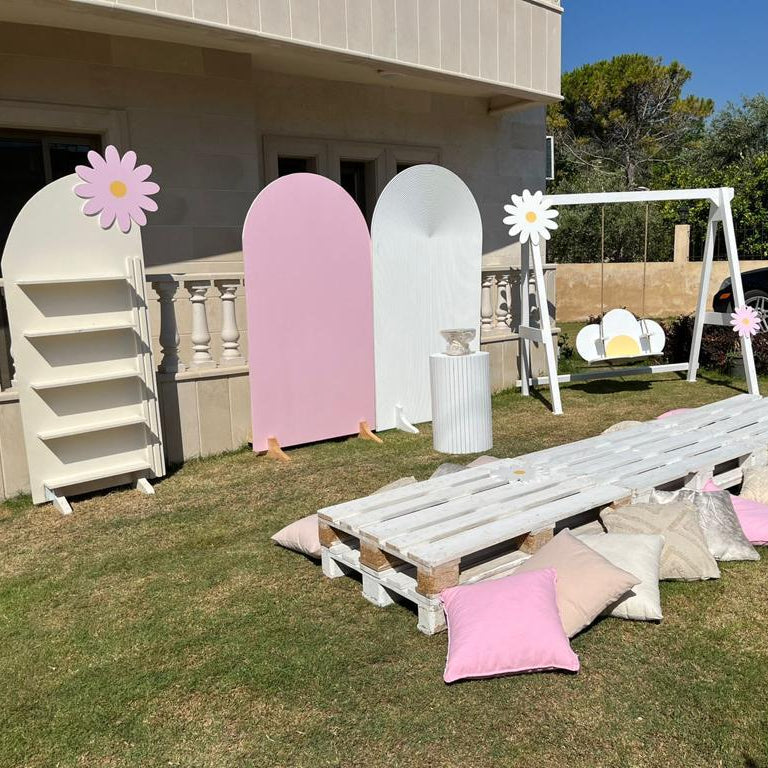 Outdoor setting with decorative items including a white pallet, pink and white boards, and pillows on grass.