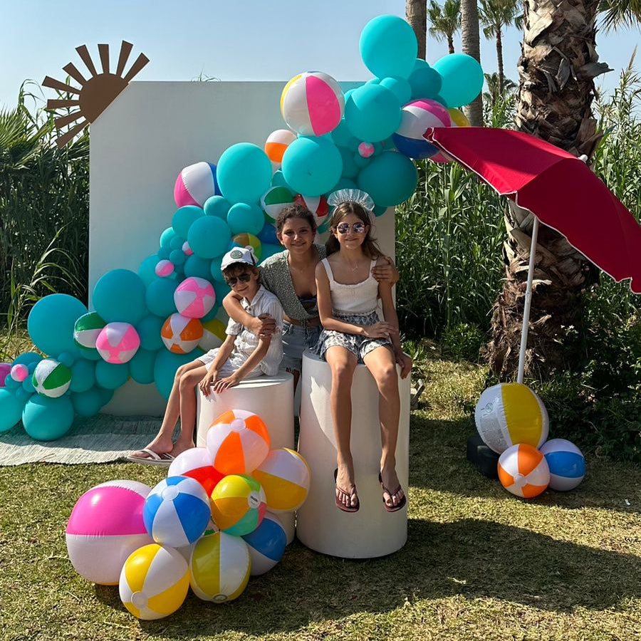 Two women sitting on stools with colorful balloons and a palm tree in the background