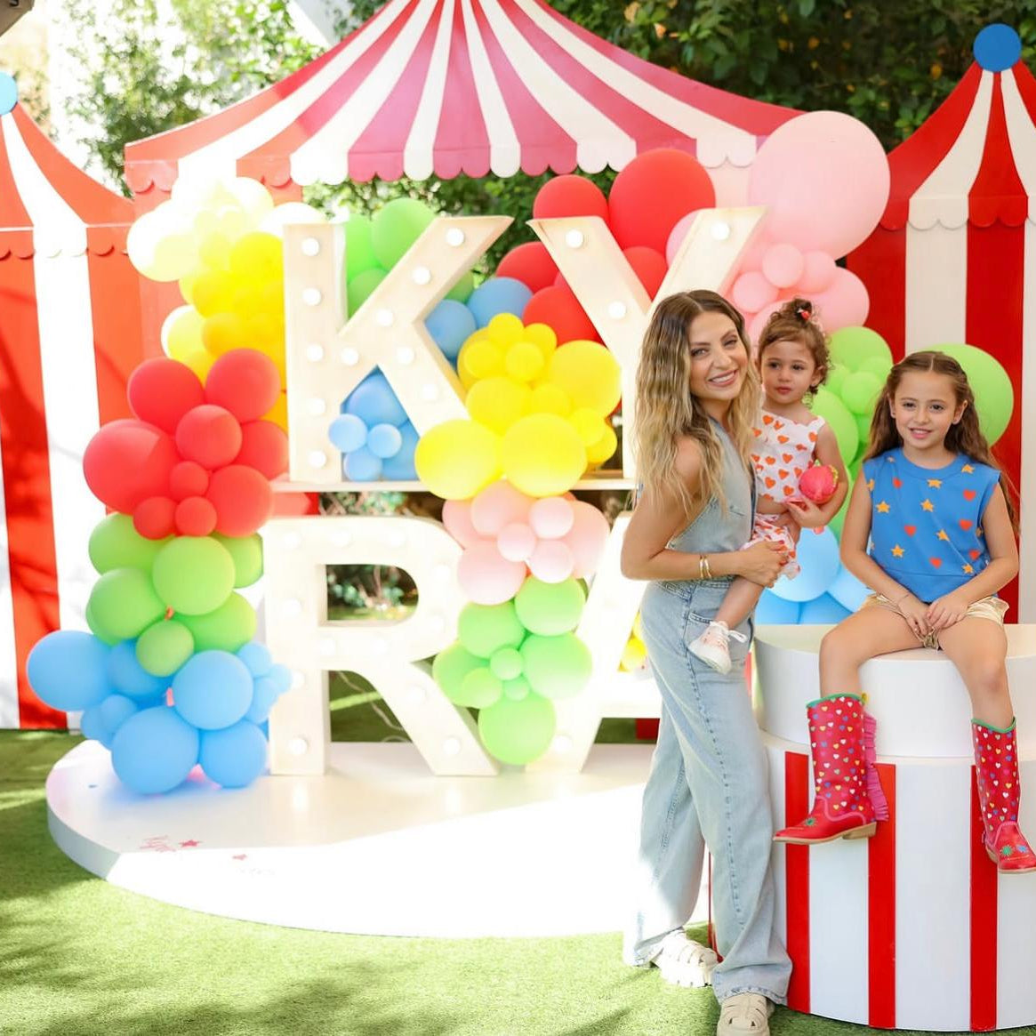 Woman with two children in front of a colorful balloon display and striped tent.