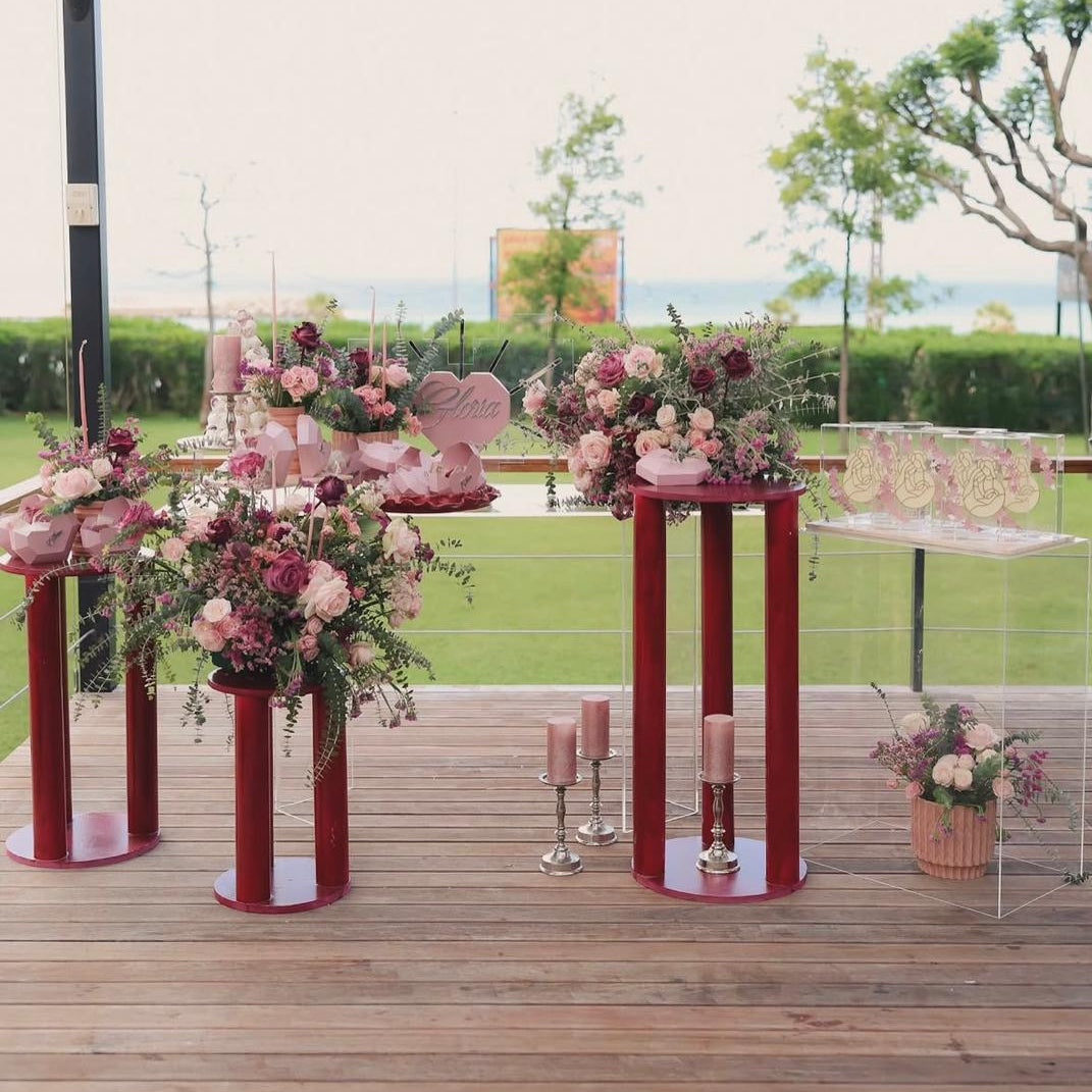 A styled outdoor dessert or event table setup on a wooden deck, featuring pink and burgundy floral arrangements, pink candles, and pink decorative stands, with a blurred green lawn and trees in the background.