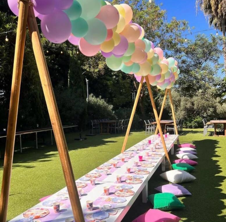 Outdoor setting with a long table decorated with pastel balloons and tableware, surrounded by greenery.