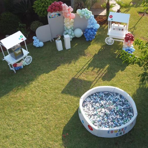 ball pit for kids'birthday with colorful balls on grass, surrounded by decorative flowers and small tables.