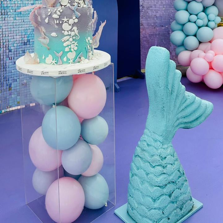 Decorative setup with balloons, a cake, and a mermaid tail on a purple floor.