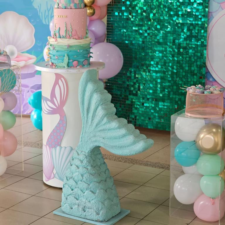 Mermaid-themed birthday party with a cake on a mermaid tail stand, balloons, and a sequin backdrop.