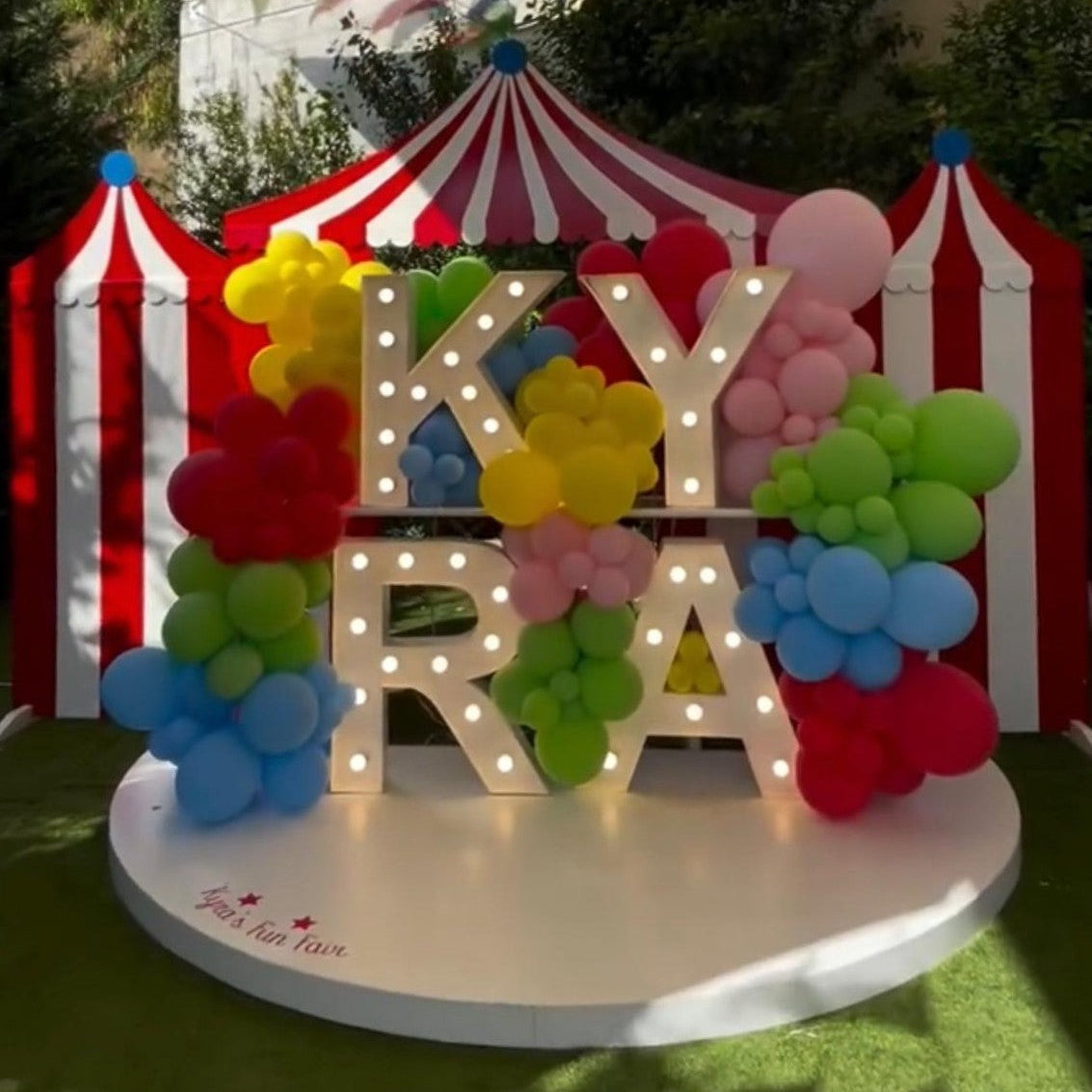Decorative setup with colorful balloons and 'KYRA' letters in front of a red and white striped tent.