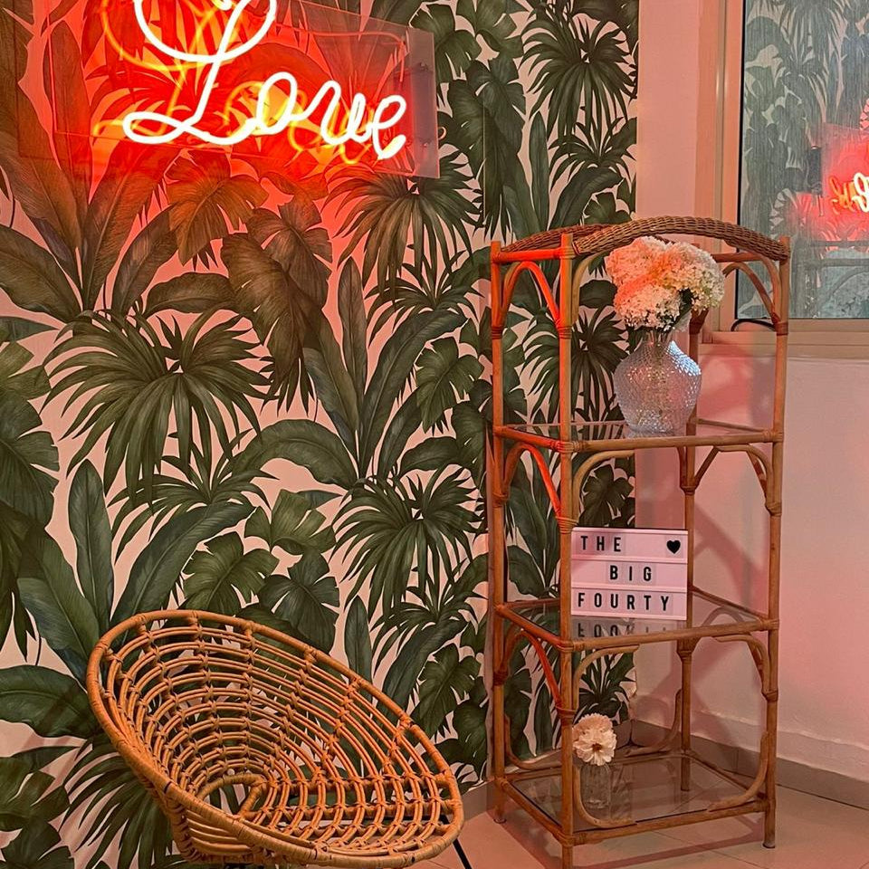 Neon 'Love' sign on a wall with tropical leaf pattern, wicker chair, and decorative shelf.