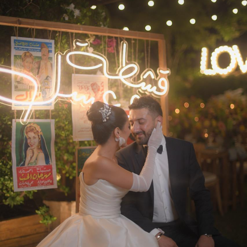 Couple in wedding attire sitting together with neon signs and posters in the background