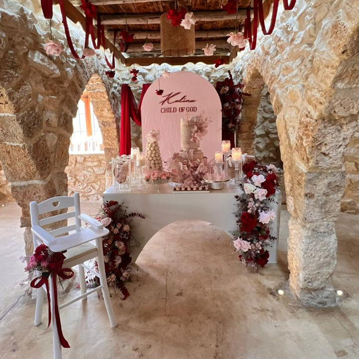 A rustic wedding reception setup in a stone hall, featuring a decorated table with a pink arch backdrop, hanging red ribbons, white chairs, and floral arrangements.