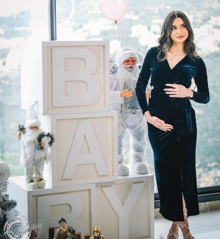 Woman in a black dress standing next to 'BABY' letter blocks with festive decorations in the background.