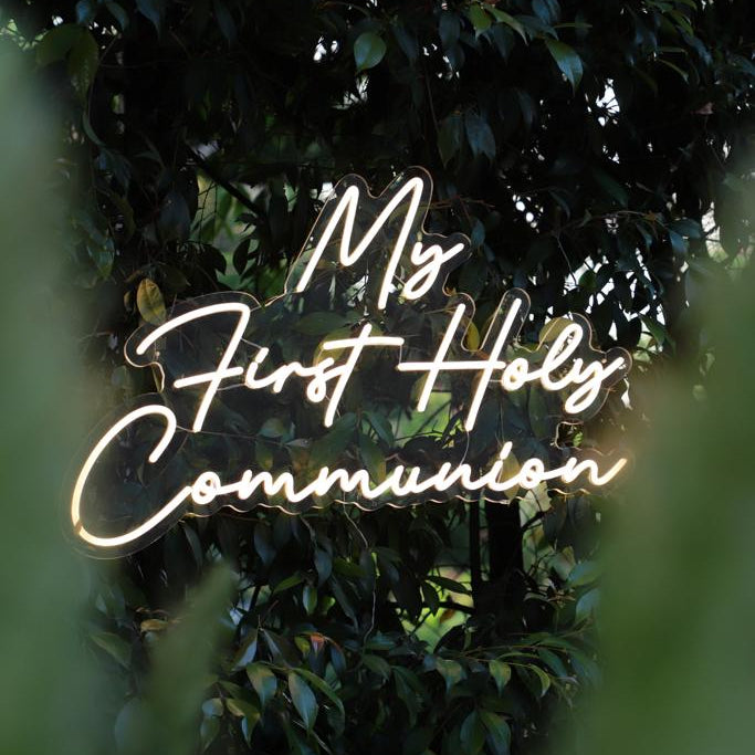 A glowing white neon-style sign reading 'My First Holy Communion' centered against a dark, leafy outdoor background with blurred green foreground elements.