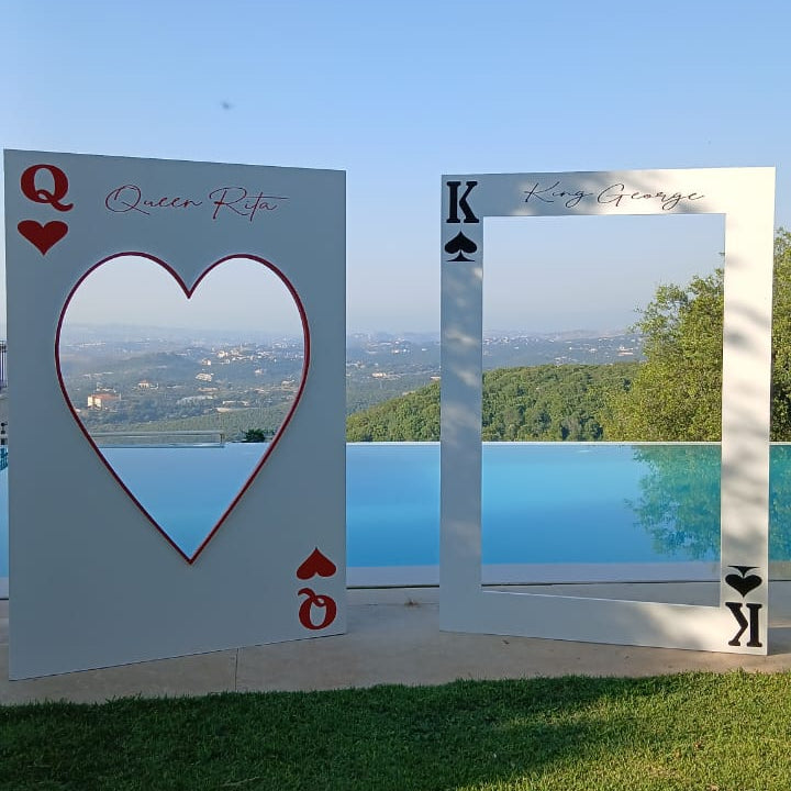 Two large white playing card photo frames on a grassy lawn by an infinity pool, with a heart-shaped cutout on the left and a king frame on the right, set against a blue sky and distant landscape.