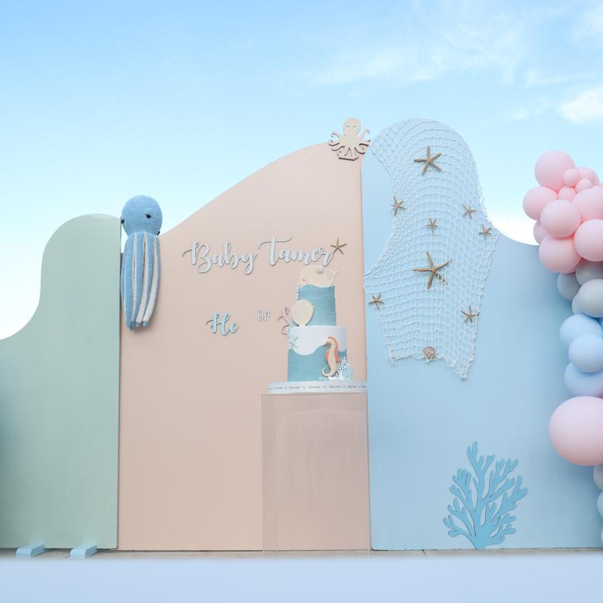 A pastel baby shower backdrop setup with a central arch featuring the words 'Baby Shower' and a small angel decoration, flanked by blue and pink balloon garlands against a light blue sky.