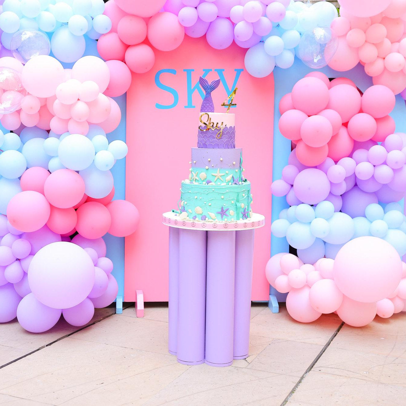 Pastel-colored balloon arch with a small cake on a stand in the center.
