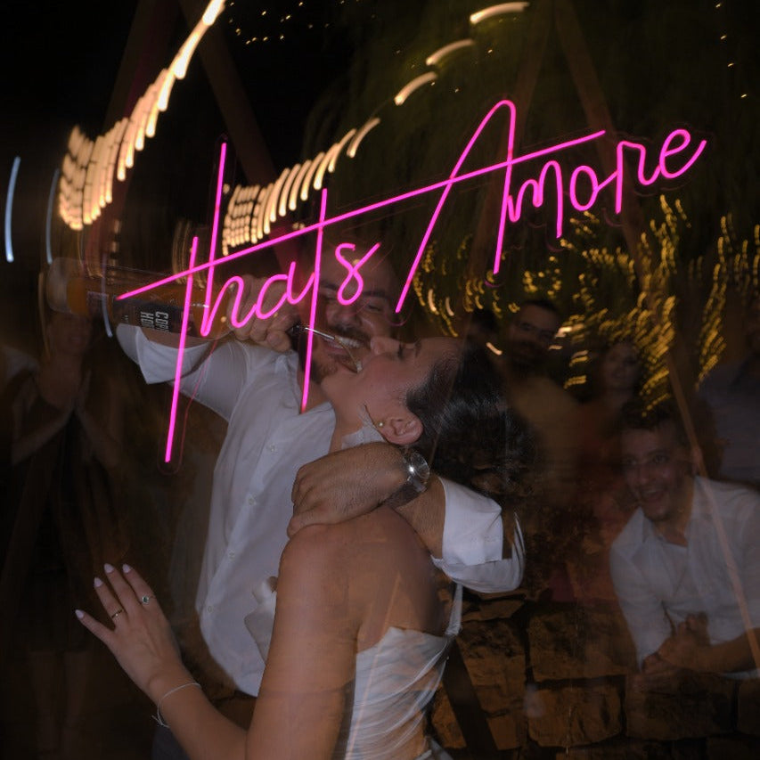 A bride in a white wedding dress and a groom dancing closely at night, with a bright pink neon sign reading 'that's Amore' in the background and warm light trails.
